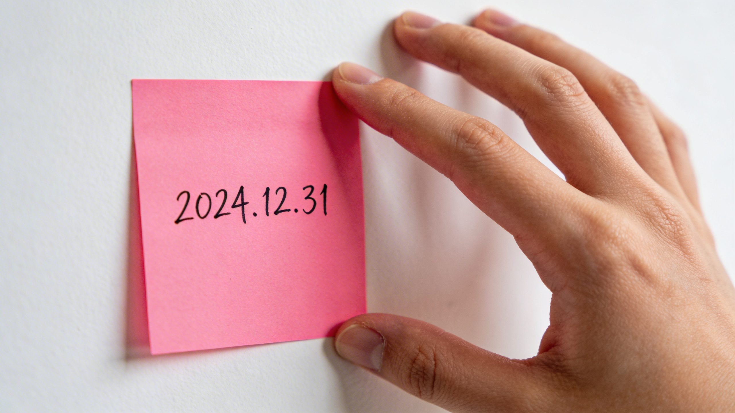 Closeup of a person’s hand placing a bright pink sticky note on a white wall with a simple payoff date written