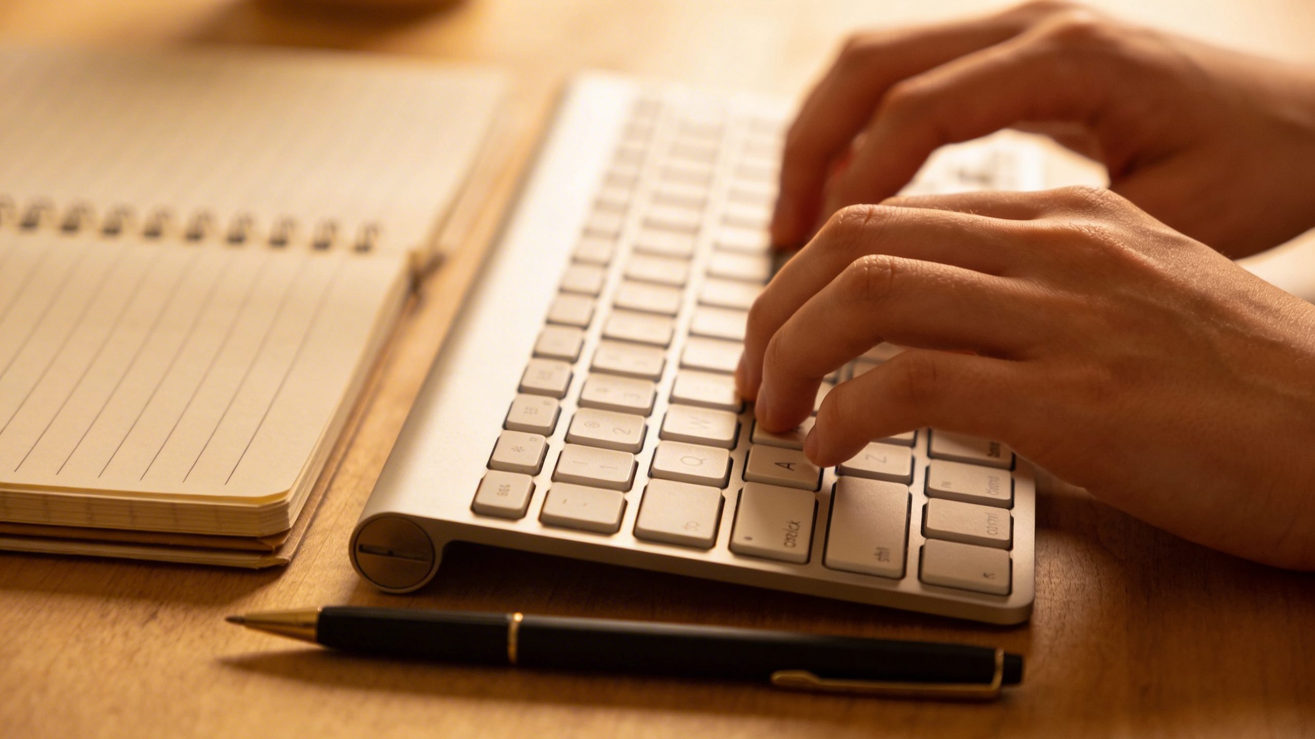 Closeup of a person typing on a keyboard with a notebook and pen beside